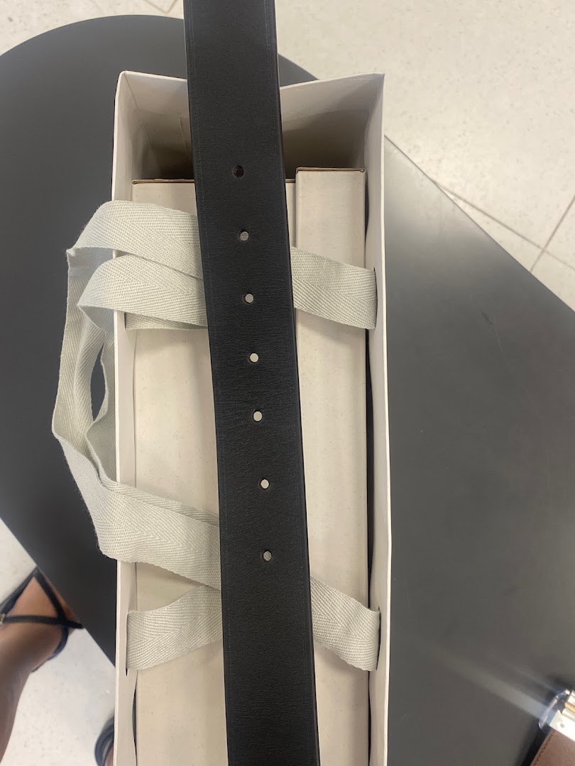 A black leather belt is presented in a white box with light grey straps.