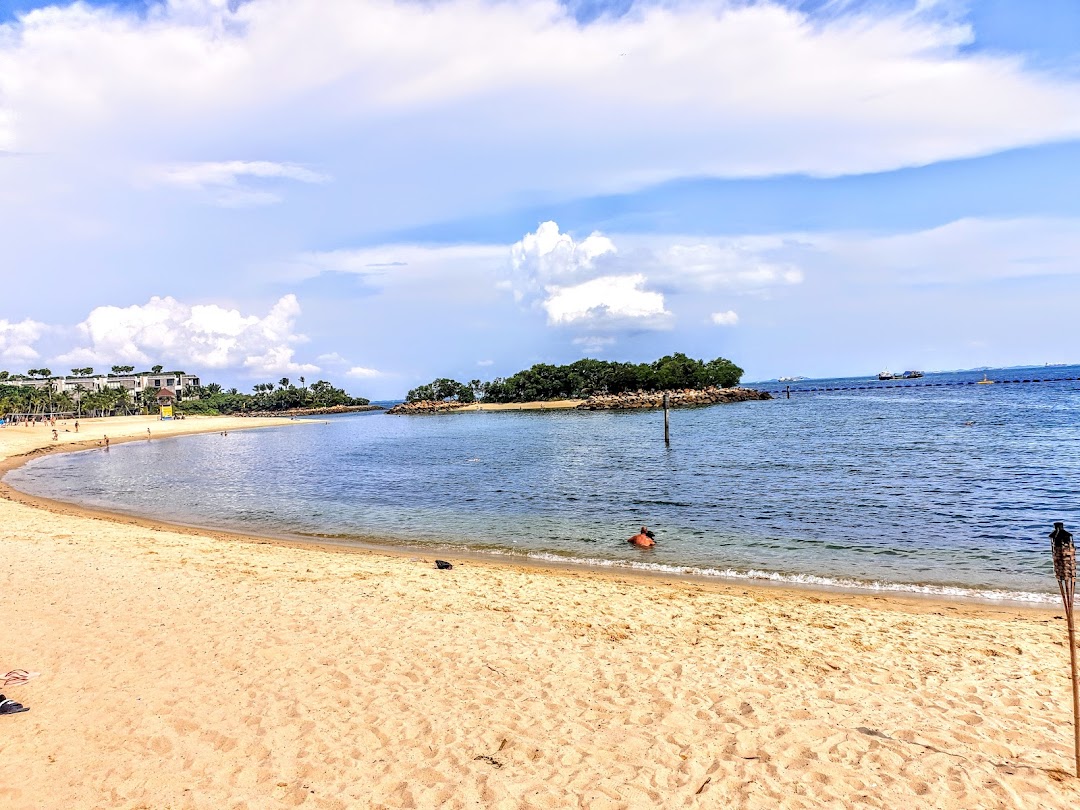 A scenic view of Tanjong Beach Club with a sandy beach, calm blue water, and distant buildings under a cloudy sky.