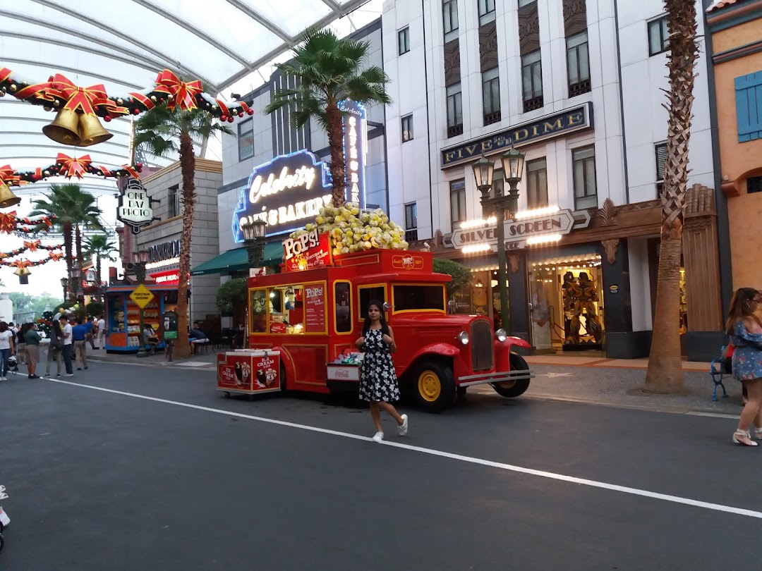 A red food truck selling popcorn and drinks on a street with shops and palm trees.